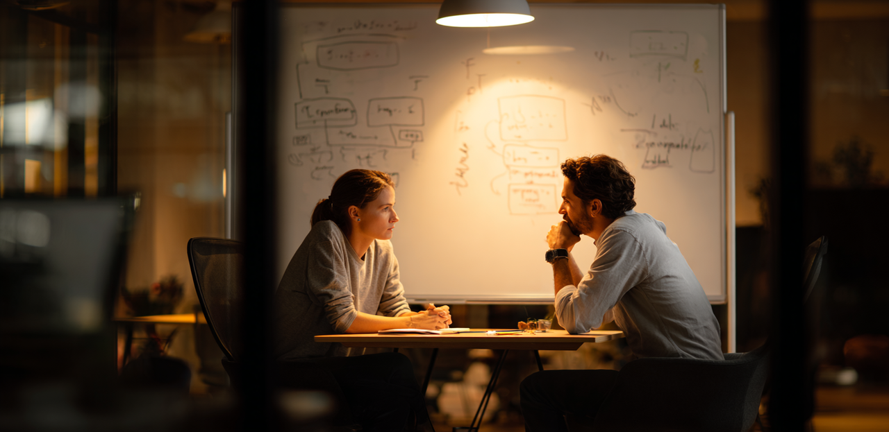Two professionals in conversation at a whiteboard covered in user flow diagrams, representing the product discovery process before writing code.